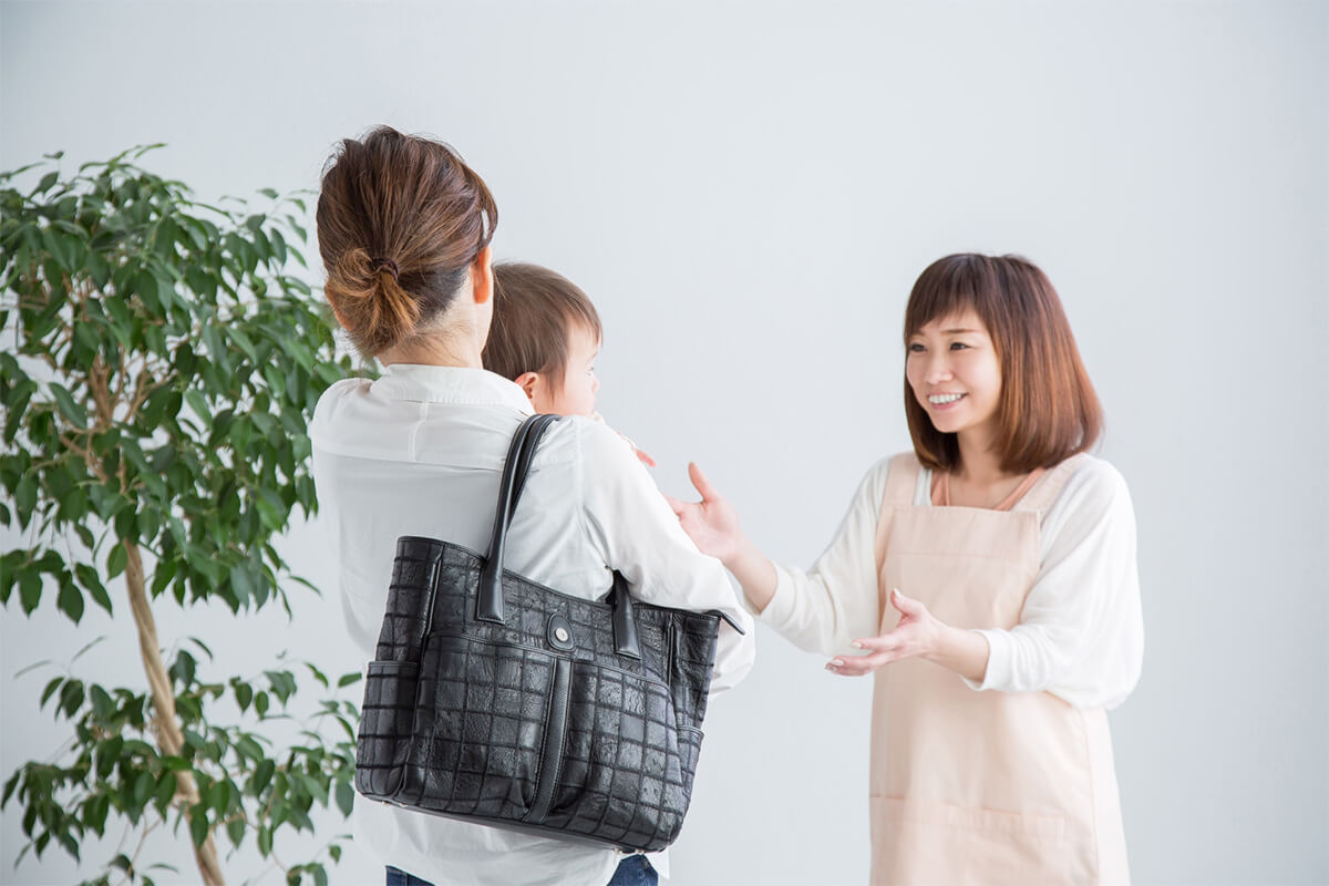 薄いピンク色のエプロンをした女性が幼児を受け取ろうとしている。手前には幼児を抱っこし、黒いビジネスバッグを肩からかけている女性。