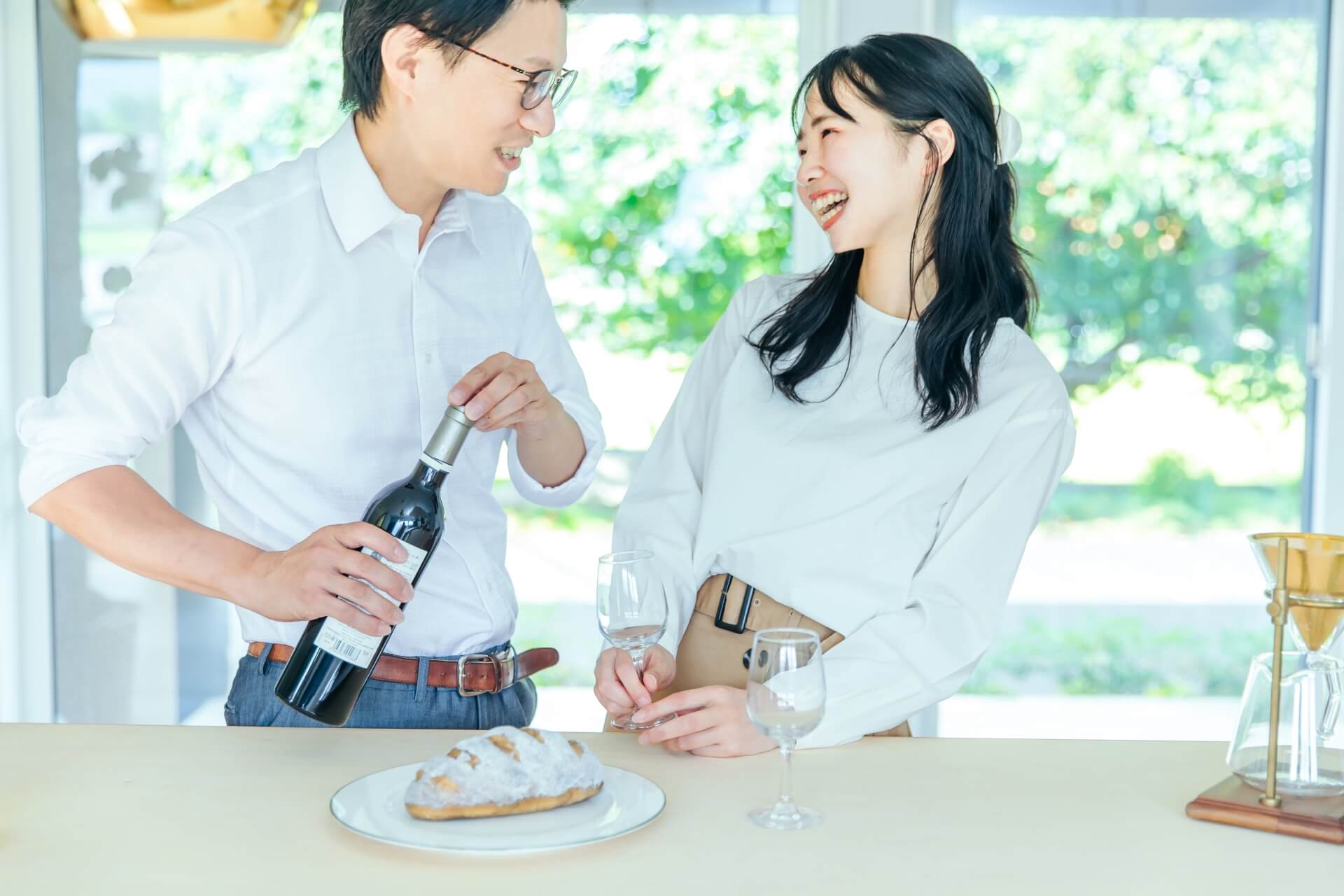 ワインボトルを持っている男性とグラスを持っている女性がカウンターの奥で談笑している。二人とも白いトップス。カウンターにはグラスがひとつとパンがおいてある。
