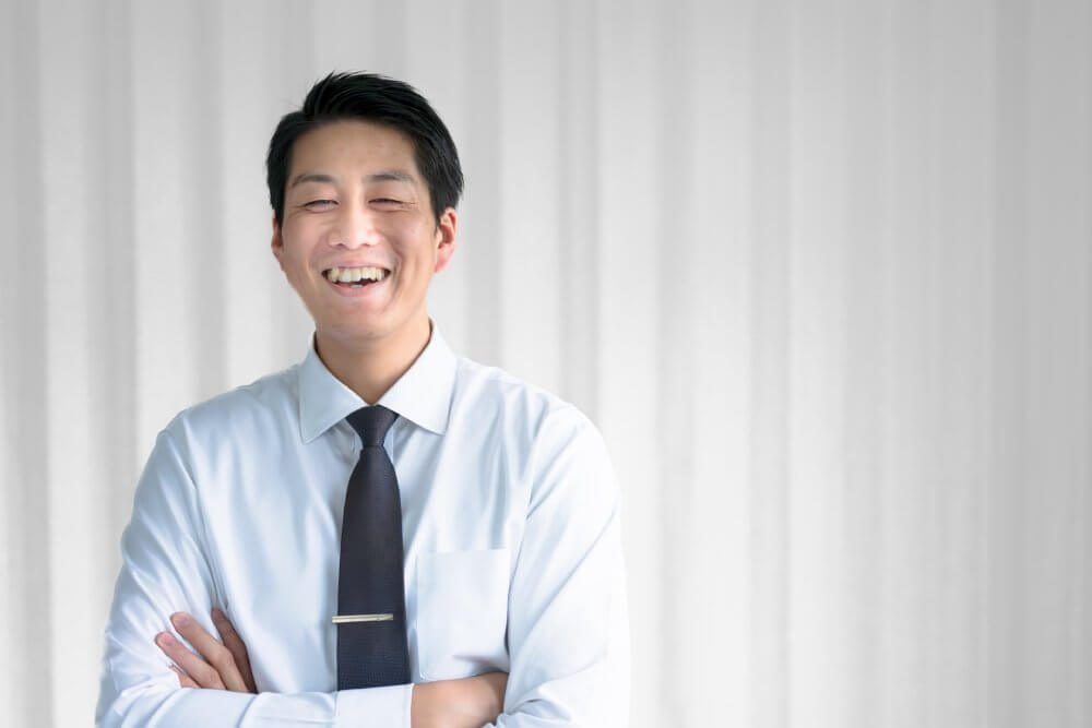 白いシャツにネクタイをし、腕組みをしている男性。満面の笑み。