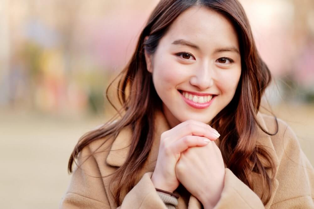 ベージュのコートを着て左手のこぶしを右手で包み、笑顔で正面を見ている女性