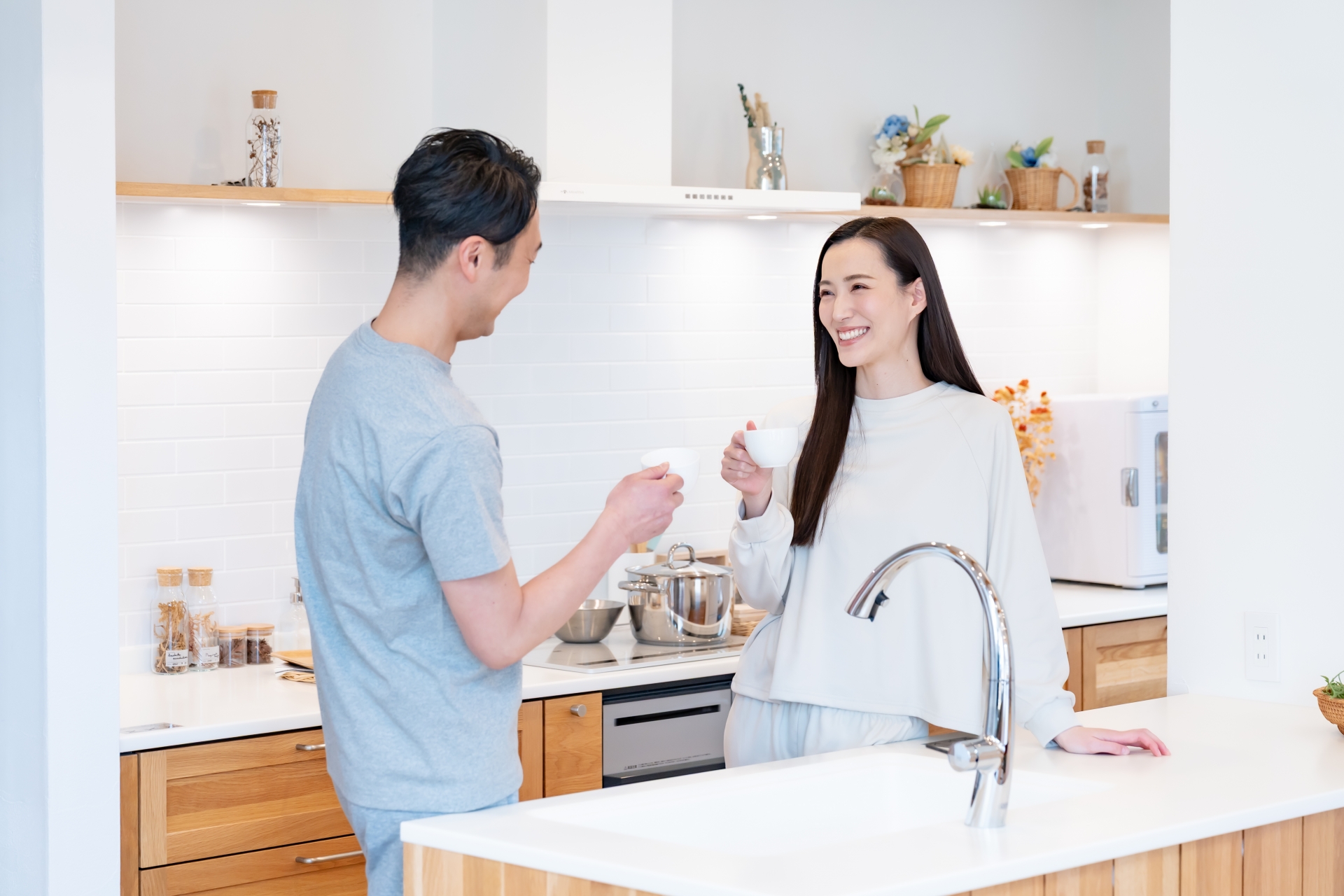 キッチンで談笑しながらコーヒーを飲んでいる夫婦。二人とも白やグレーの部屋着を着ている。