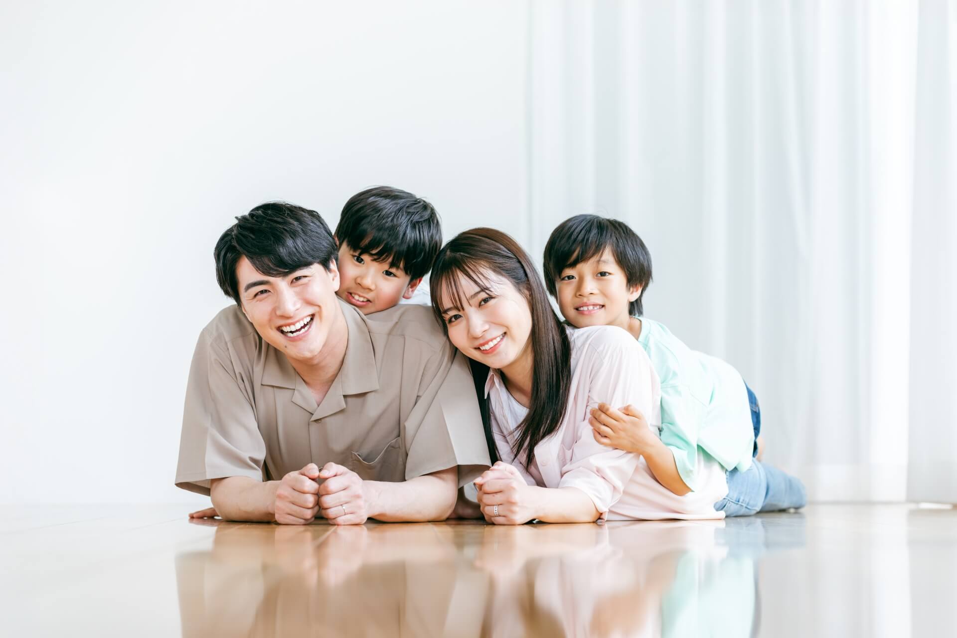 リビングに寝そべって正面に笑顔を向けている4人家族。若い両親の背中の上に男の子が2人乗っている。