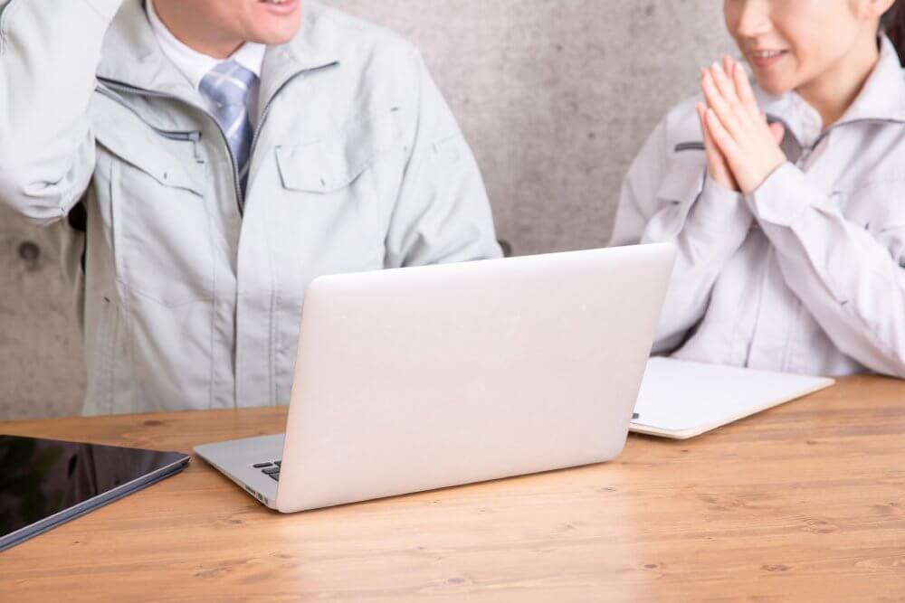 ノートパソコンの前にいる作業着の男女。女性が手を合わせて笑顔。男性は右手を頭の方に向けて照れているようなしぐさ。