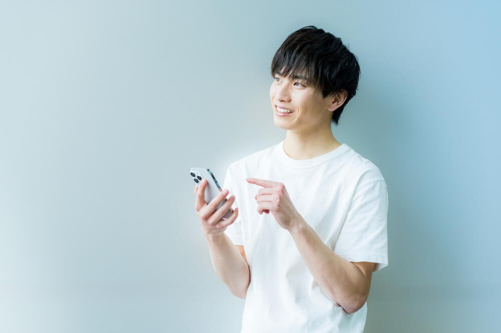 31944356_m 白いTシャツの男性が左上に視線を向けながらスマホを触ろうとしているところ。