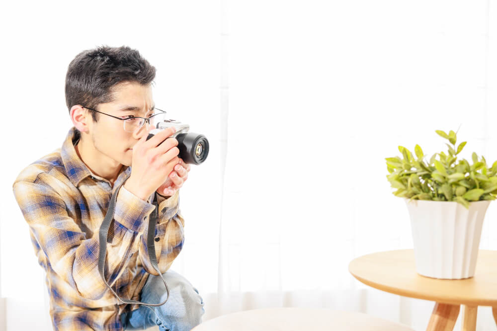 室内でミニテーブルの上の観葉植物をミラーレスカメラで撮影する男性。男性はメガネをかけており、黄色と青のチェック柄のシャツを着てしゃがんだ体勢