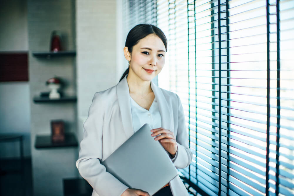 ブラインドの窓際に立つ、ベージュのジャケットを羽織りノートパソコンを小脇に抱える女性。すっきりとまとめた髪型でほほ笑んでいる