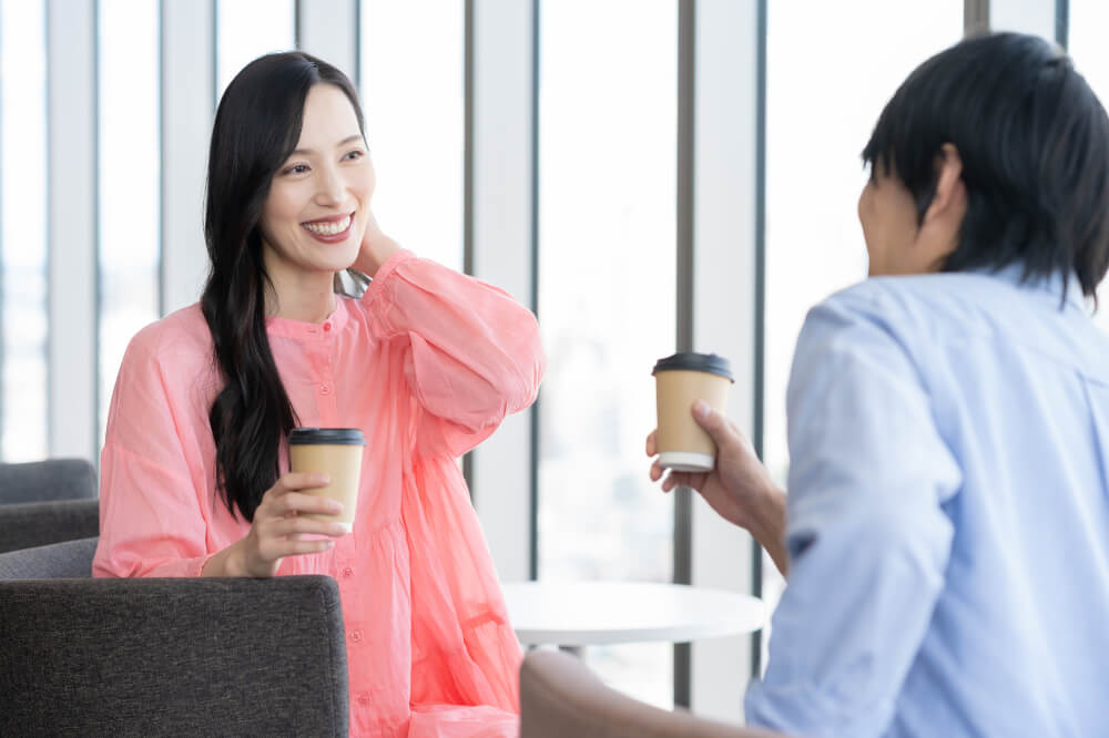ピンクのシフォンブラウスを着たロングヘアの女性、右手にコーヒーカップを持ち、笑顔で左手で髪を耳に書けるようなしぐさをしている。向かいには水色のシャツを着た男性が座っており、男性もコーヒーカップを持っている