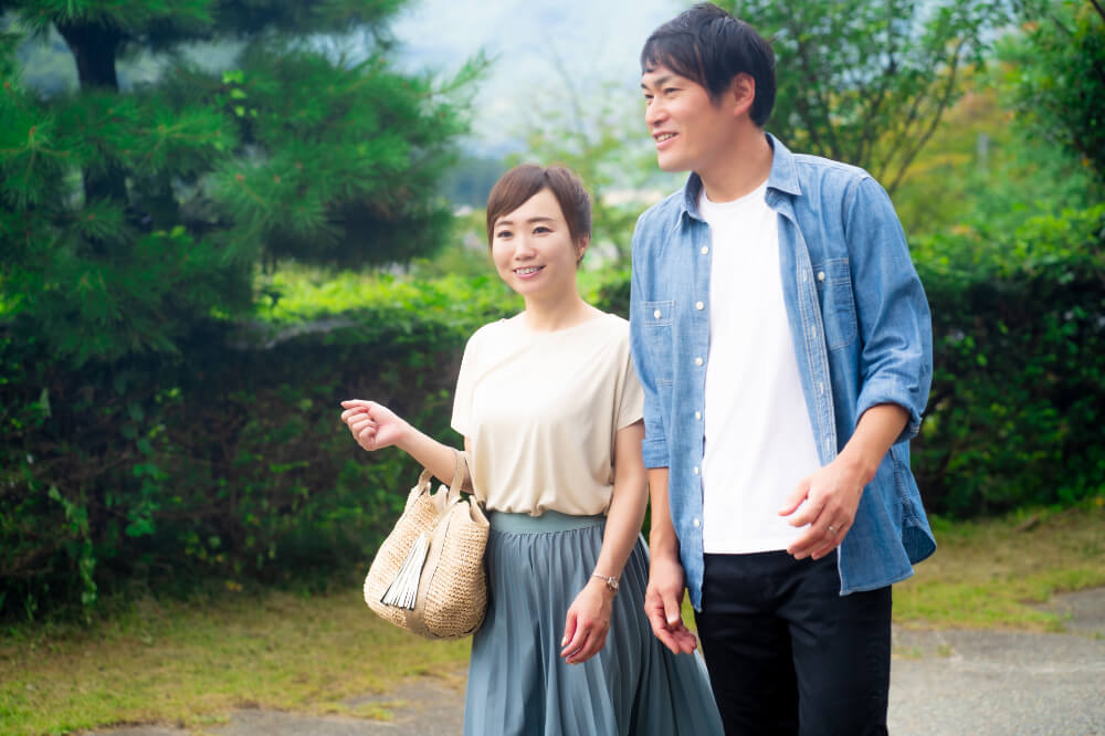 日本庭園のようなところを歩く男女。男性はデニムシャツに黒いパンツ、女性はベージュのTシャツに青っぽいグレーのスカート。二人とも笑顔で歩いている。