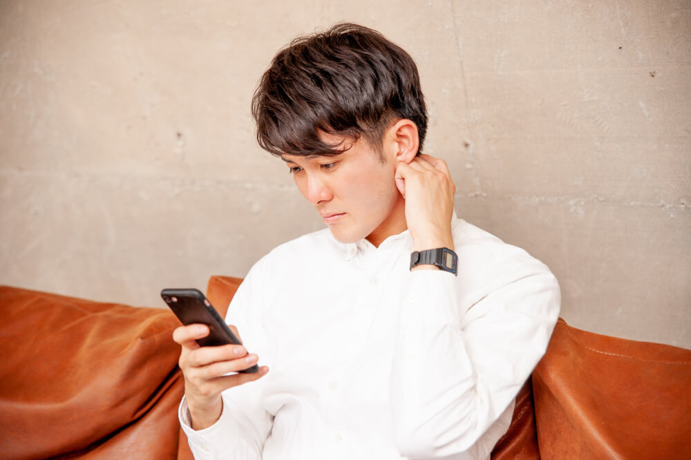オレンジっぽい皮のソファに座る白いシャツを着た男性。スマホを見ながら、すこし困ったような表情