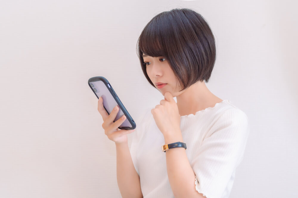 スマホを見る女性 白いトップスを着たボブヘアの女性がスマホを見て少し悩んでいるような表情。