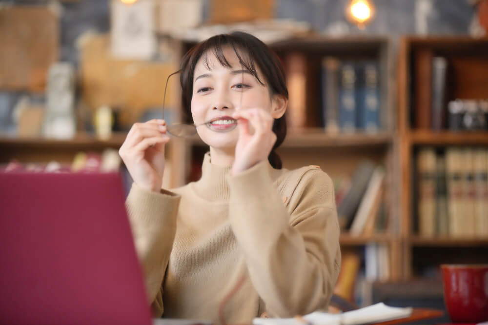えんじ色のノートパソコンの前に座る女性。ベージュ色のハイネックニットを着ている。背景には本棚。両手で眼鏡をはずしているところ。