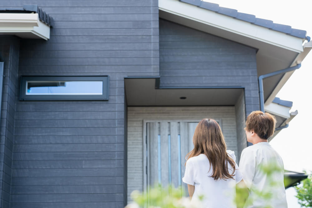 マイホームを眺める夫婦 黒っぽい外壁の新築一戸建ての前に白いトップスを着た若い夫婦が立っている