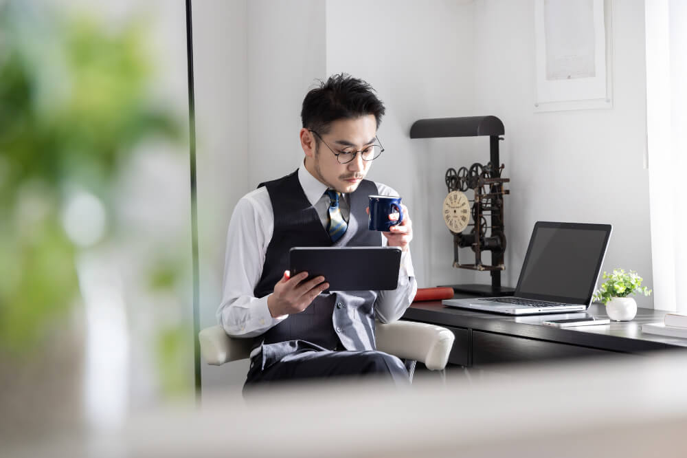 ノートパソコンが置いてある窓辺のデスク。ワイシャツにベストを着用したメガネをかけた男性がコーヒーを片手に座っている。もう片方の手で持ったタブレットの画面を見ている。