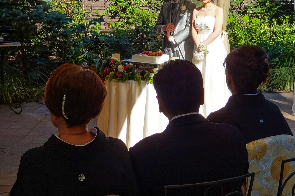 奥にはウェディングケーキ、ケーキの後ろに新郎新婦が立っている。手前の椅子に、親族らしき着物を着た女性が2人と、男性が一人、椅子に座っている。