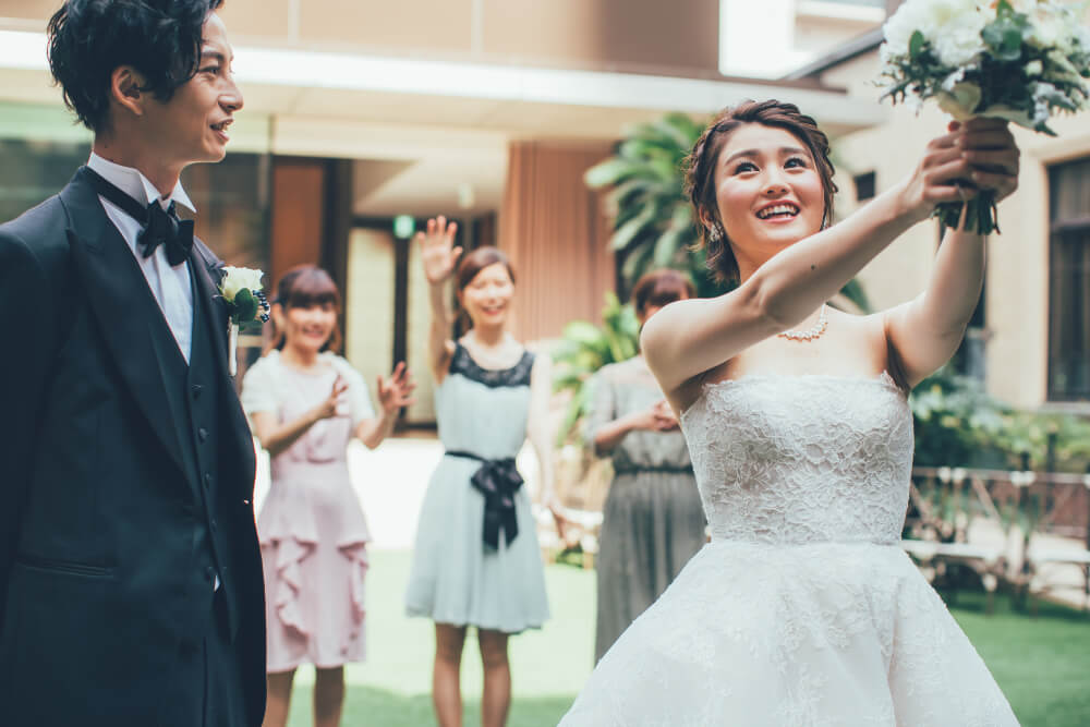 ガーデンウェディング会場で今からブーケを投げようとしている新婦と、それを見守る新郎。背景にはブーケをキャッチしようとしているゲストたち