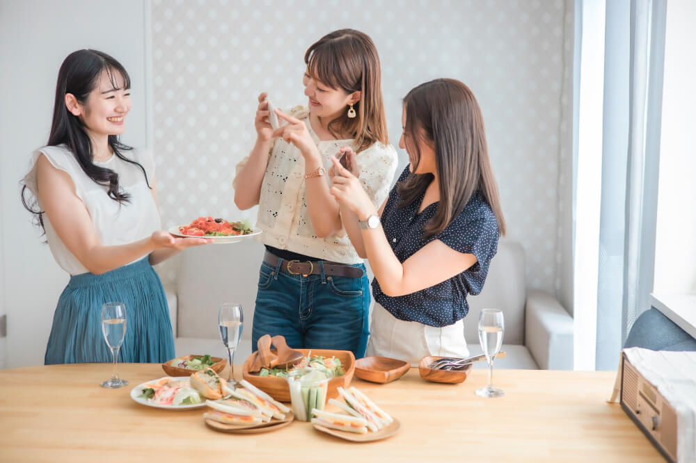サンドイッチとサラダでホームパーティをしている女性三人。ひとりの女性が持っているサラダの写真を撮っているところ