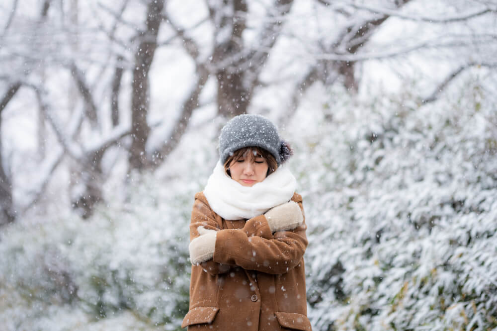 雪景色の山のようなところで、防寒着で両腕で自分を抱きしめるようなポーズで凍えている女性。