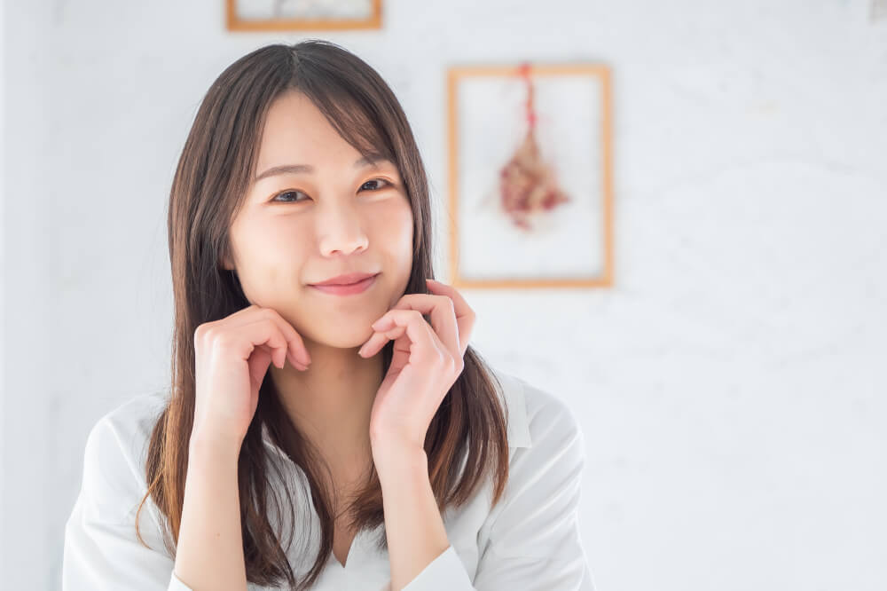 ほほ笑む女性 こちらを向いてほほ笑んでいる女性。両手を頬に添えている。背景にはアートが飾ってある白い壁。