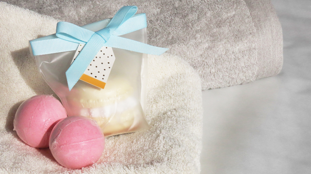 白いタオルの上に置かれたギフト。水色のリボンがかかった袋に、バスボムが入っている。袋の外に、ピンク色のバスボムも二つ置いてある