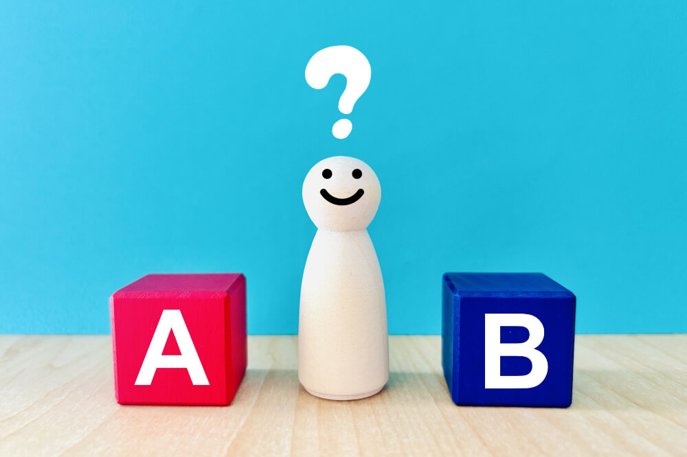 AかBか 水色背景にAと印字された赤いブロックとBと印字された青いブロック、その間に白い木の人形が置いてある。人形の上にはてなマークが浮いている