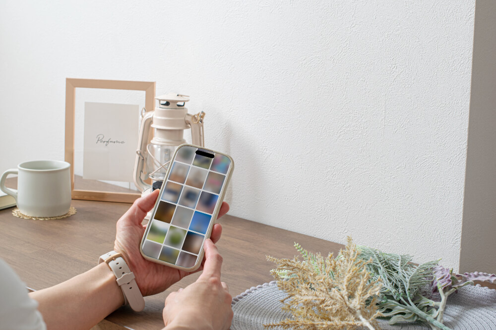 白い壁側にくっついた木のデスクにコーヒーカップや写真フレーム、インテリアランプが置いてある。手前にはスマホを持つ女性の手。カメラロールを見ている。