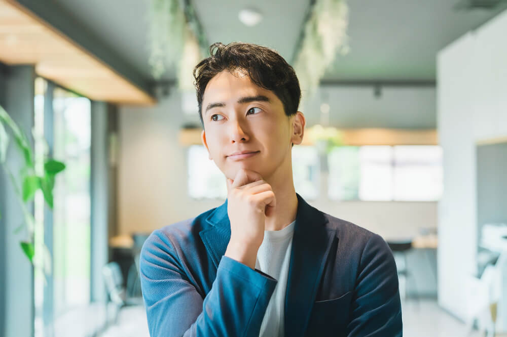 前向きに考える男性 カフェのような背景に、紺色のジャケットの男性が右手を顎に添えて考え事をしている。ほほ笑んでいるので前向きなことを考えている様子。
