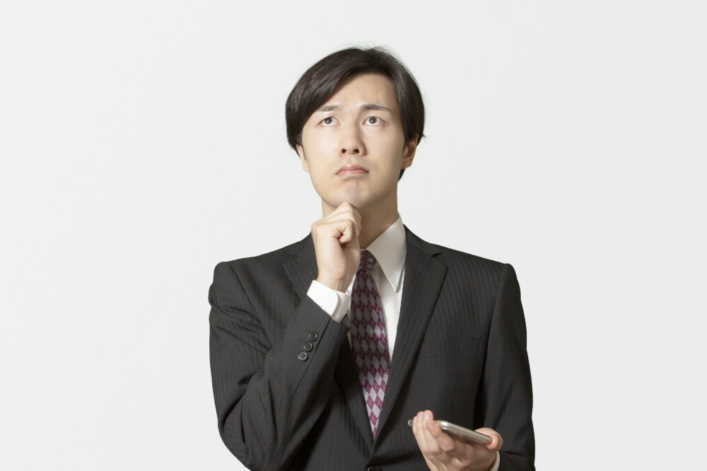 悩む男性 スマホを左手に持ち、右手を顎に添えて、目線を上にしながら悩む男性。アーガイル柄ネクタイに薄いストライプ模様の入った黒いスーツを着ている。