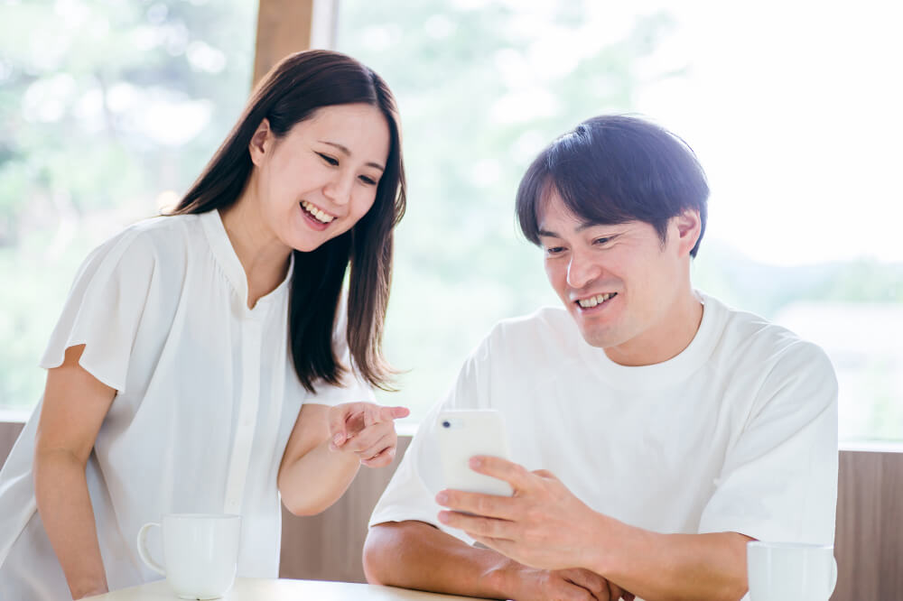 白いトップスを着た男女。大きな窓のある室内。男性は席についていて手元にスマホを持っている。横に立っている女性も男性のスマホを指さして見ながら笑っている。