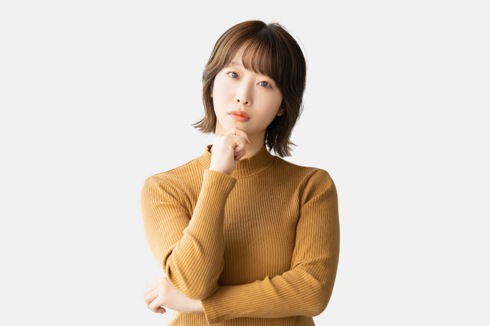 マスタード色のタートルニットを着たショートカットの女性。腕組みをして右手をあごに添えて小首をかしげている