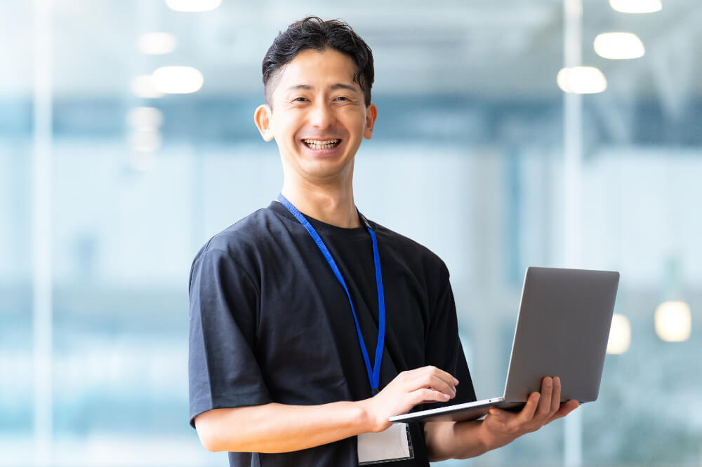 黒いTシャツに、青い紐のネームホルダーを首から下げた笑顔の男性。ノートパソコンを持っている