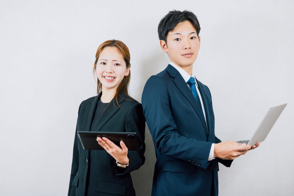 スーツ、ジャケットを着た女性と男性が笑顔で背中合わせに立っている。女性はタブレット男性はノートパソコンを持っている